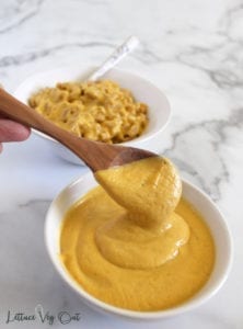 Wooden spoon scooping vegan cheese sauce with pasta in bowl behind