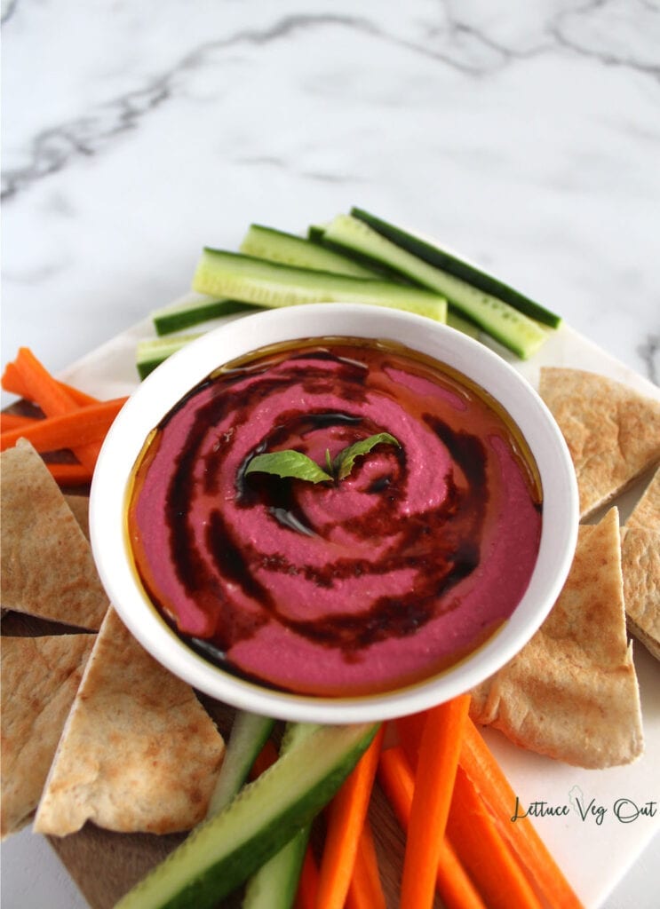 White bowl of vegan beetroot dip topped with olive oil and balsamic on platter with pita, carrot and cucumber surrounding the dip