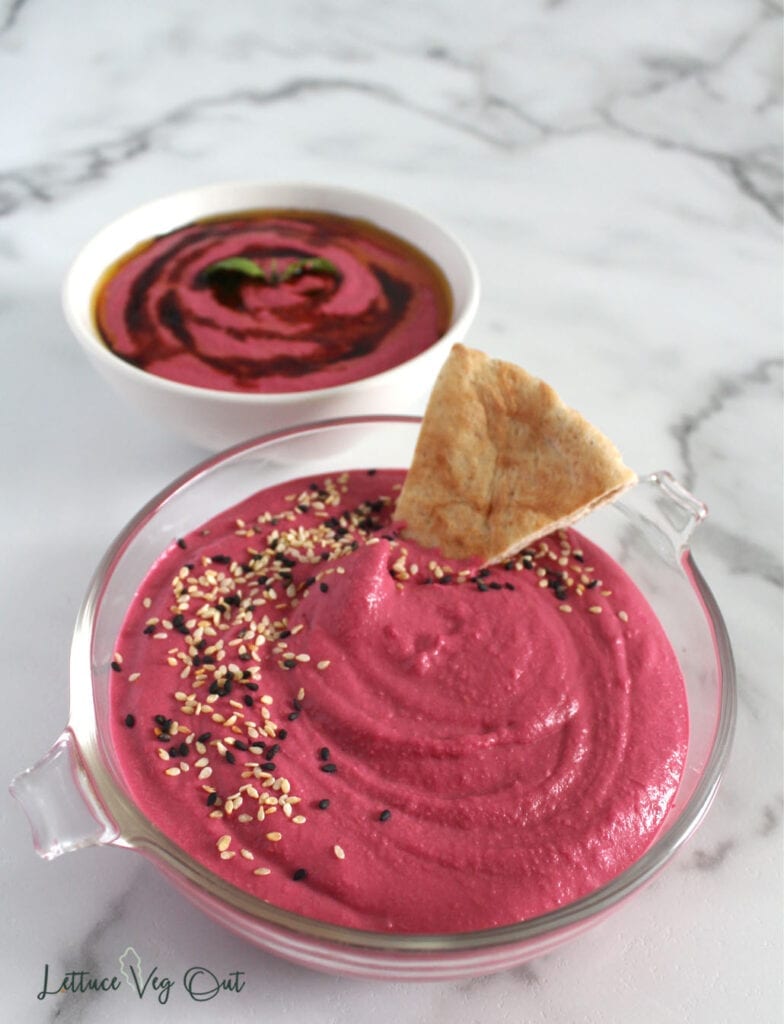 Glass bowl of pink beet dip with pita bread in dip and back white bowl of dip topped with balsamic on marble background