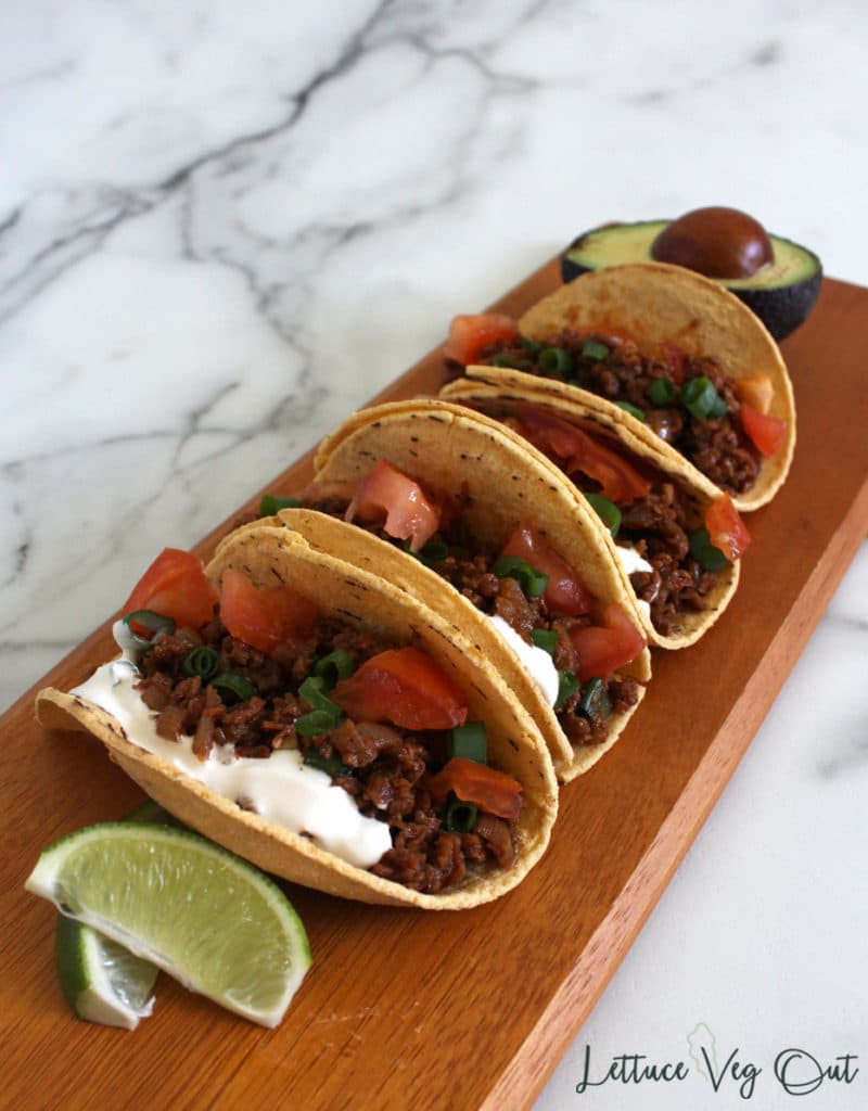 Row of vegan TVP tacos on wooden cutting board with sour cream and tomato