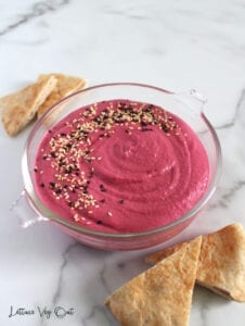 Glass dish of pink vegan beetroot dip with half circle sprinkle of white and black sesame seeds and a couple triangles of pita bread around the dip