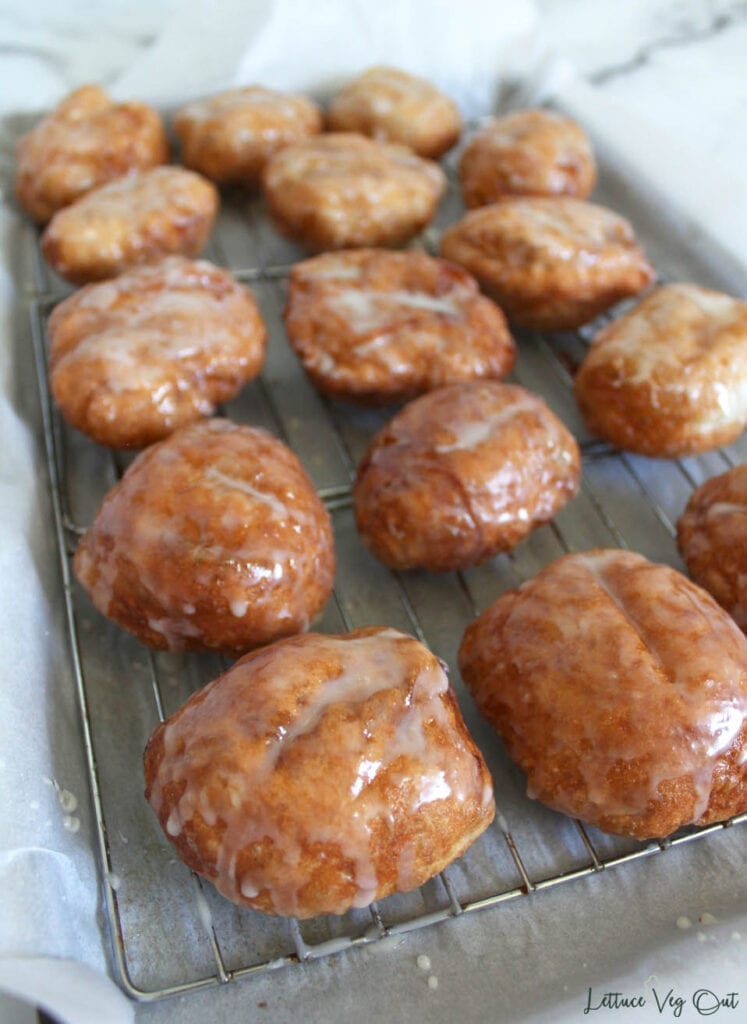 Eggless apple fritters on a cooling rack above a parchment paper covered baking tray, coated in glaze