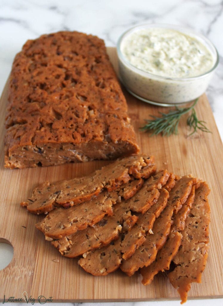 Seitan gyro meat, half of it sliced, on wooden cutting board with glass jar of sour cream sauce and fresh dill on the back right of the board
