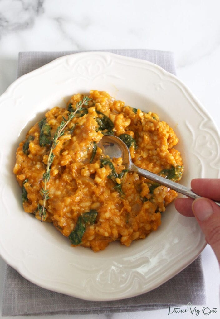 Hand holding spoon and scooping bite of creamy pumpkin risotto from a white plate resting on grey towel set upon marble counter