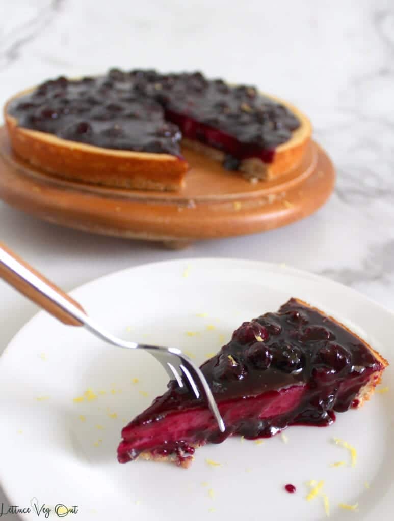 Fork pressing into first bite of blueberry cheesecake slice sitting on white plate with cheesecake in background