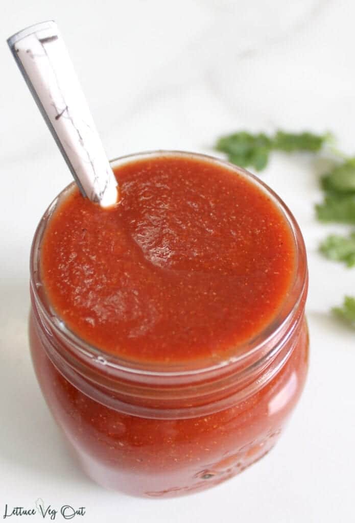 Small glass jar full of thick red vegan enchilada sauce with marble patterned spoon sitting in jar and sprig of cilantro blurred in the back right
