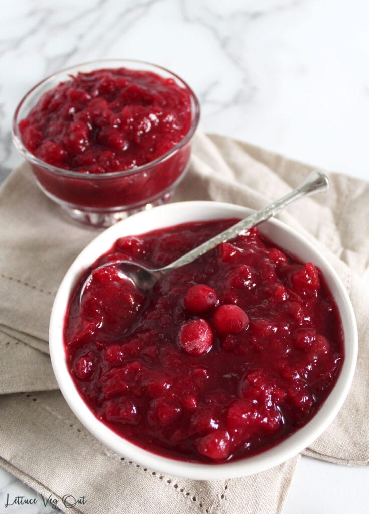Two bowls of vegan cranberry sauce - one in a glass dish and another in a white serving bowl with spoon sitting on top