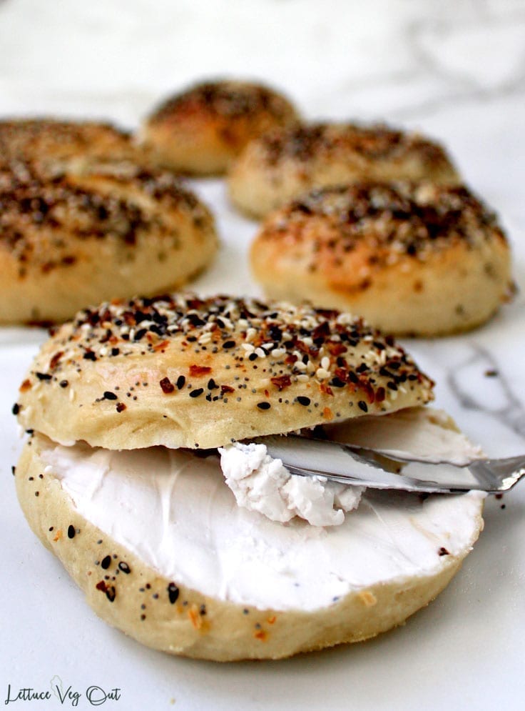 Final image of a ready-to-serve vegan everything bagel with cream cheese. Background shows other homemade bagels out of focus but still looking delicious.