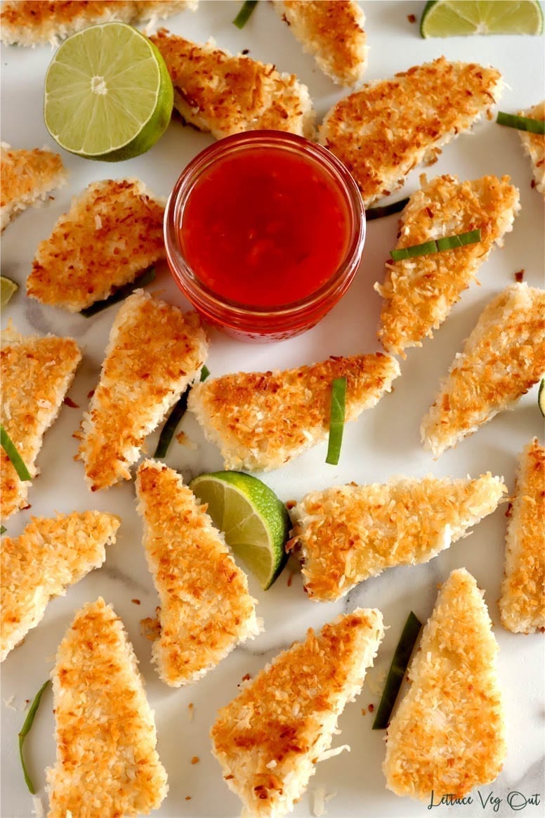 Over a dozen pieces of easy coconut crusted tofu spaced out across a granite countertop. In the middle of all these pieces is a small glass dish of sweet chili sauce