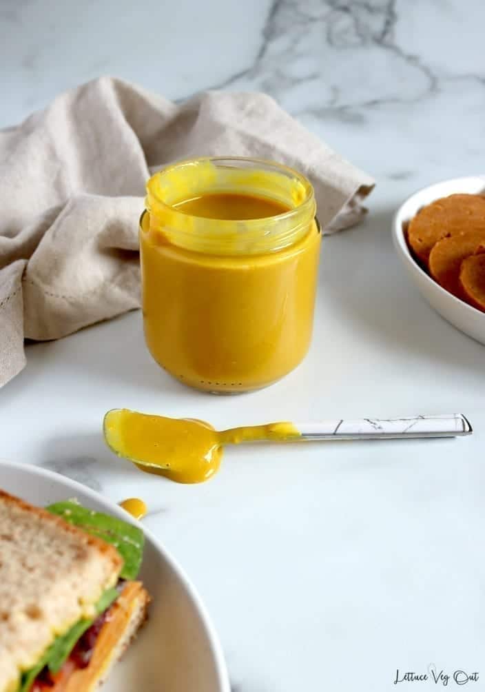 A glass jar full of homemade vegan honey mustard. In front of the jar is a small spoon that was used to spread the maple mustard onto a vegan ham sandwich