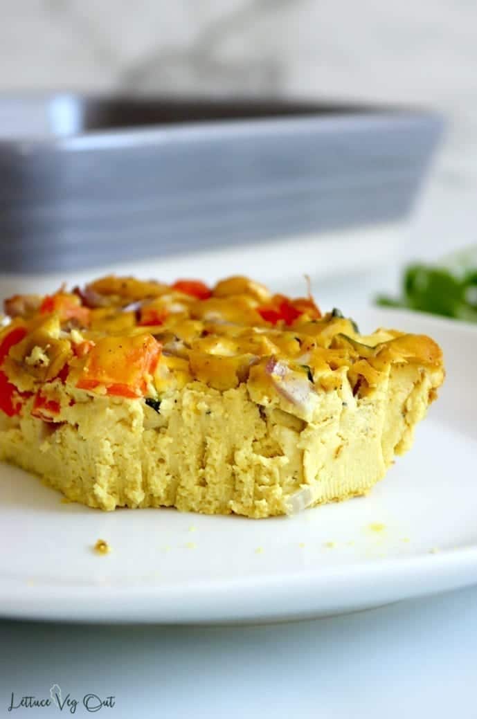 A served piece of dairy free tofu frittata on a white plate. The deep yellow color of the frittata sits in contrast to the vegetables inside, including red bell pepper, red onion and zucchini