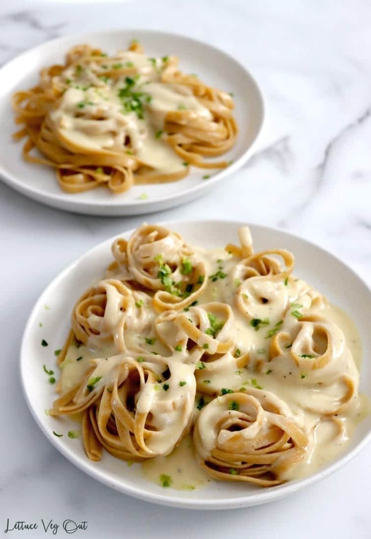 Two white plates topped with twirled pasta and covered with creamy vegan bechamel sauce and a light sprinkle of green herbs. Plate in the background partially blurred.
