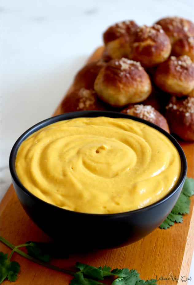 Black bowl filled with creamy beer cheese sauce sits on wood board with stack of dark brown pretzel bites on the board behind the bowl. Green garnish around bowl.