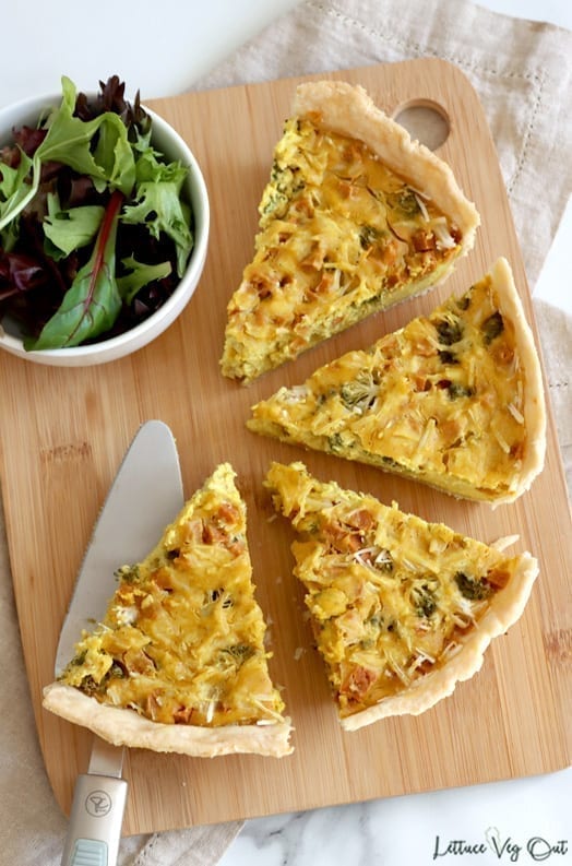 Top view of four slices of quiche sitting on wooden board. Bottom left piece resting on metal pie lifter and a small white bowl of salad greens sits to the top left.