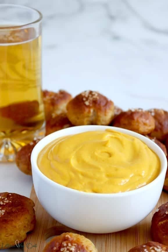 White bowl of cheese sauce on wood board surrounded with pretzel bites for dipping. A large glass beer mug, full of golden ale, sits to the back left of the sauce.