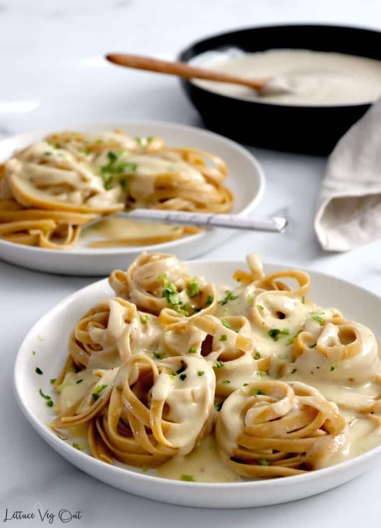 White plate with small piles of twirled linguine noodles is covered in a creamy white sauce and topped with a sprinkle of green herbs. A second plate, with a fork, pasta and sauce sits blurred behind it and further back, more blurred, is a black pan full of white sauce.