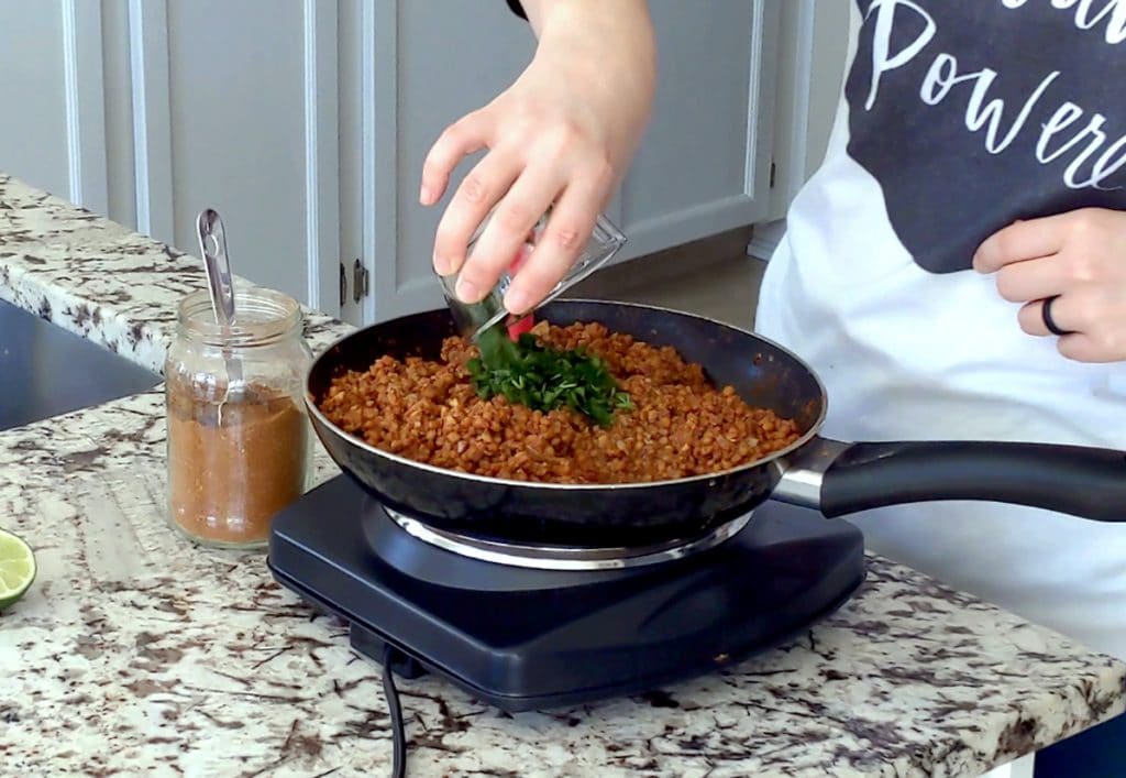 A hand pours a glass jar filled with chopped cilantro into a large black pan filled with cooked lentil and walnut taco filling. A glass jar of taco spice with a metal spoon in it sits to the left of the image.