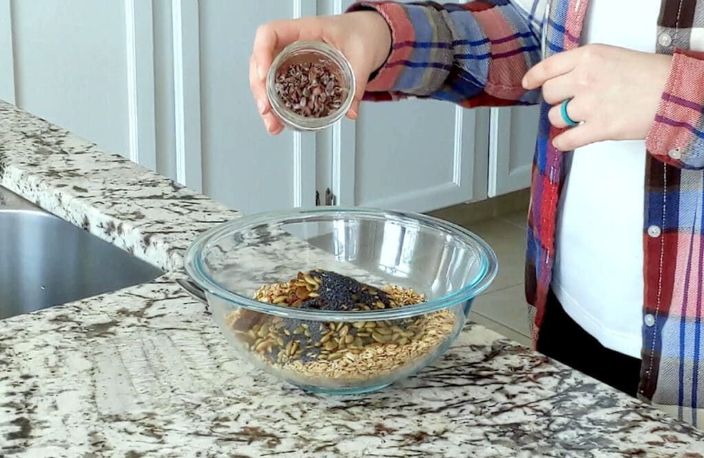 Large glass bowl on marble counter filled with oats, pumpkin seeds, poppy seeds and raisins. A hand is tipping a small glass jar of cacao nibs into the large bowl.