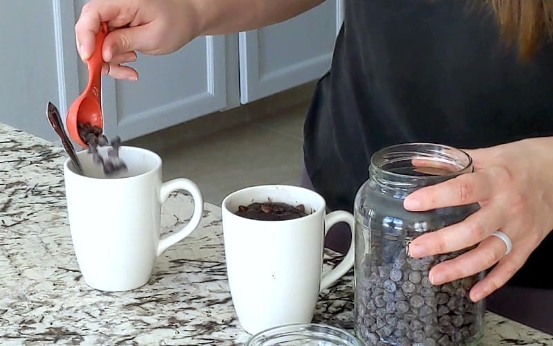 Hand holding red plastic spoon which is dumping chocolate chips into a large white mug. A second white mug sits on the marble counter to the right, full of a cooked chocolate brownie. Further right, a large glass jar filled with chocolate chips sits with a hand resting on it.