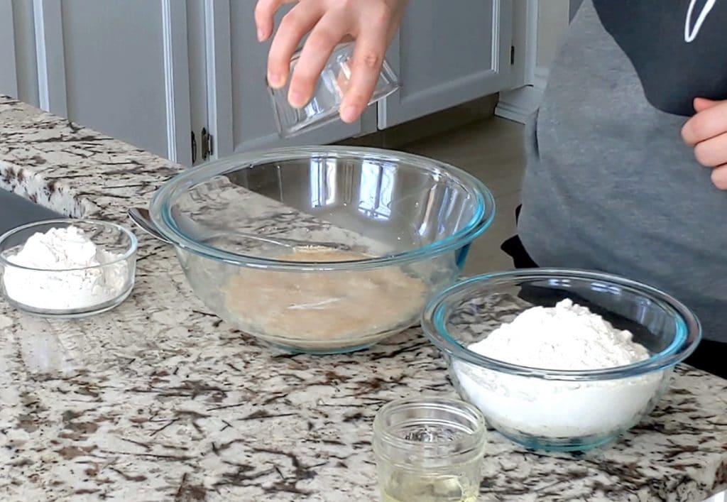 A large glass bowl filled with foamy yeast sits on a marble counter top surrounded by smaller bowls filled with flour and oil. A person holds a glass dish upside down over the large mixing bowl.
