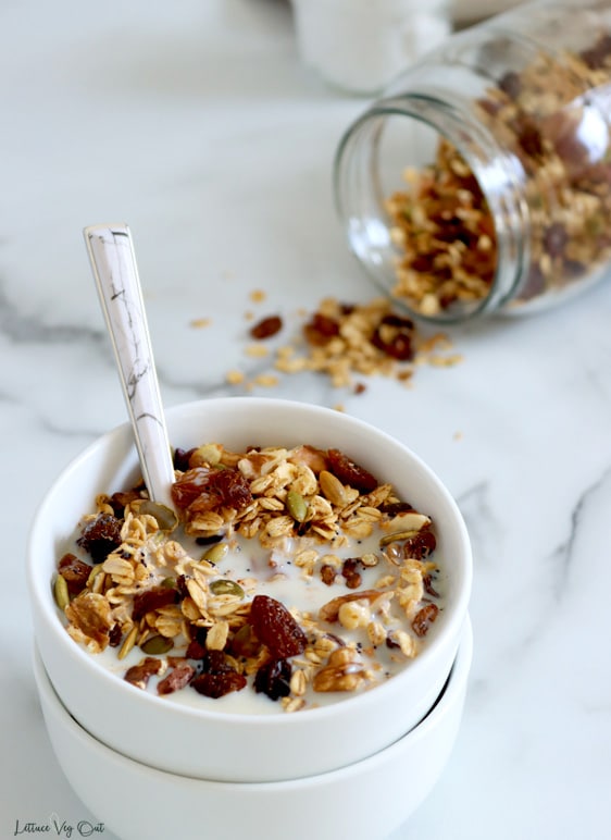 A double stack of small white bowls is filled with muesli (oats, raisins, cacao nibs and pumpkin seeds visible) with milk and a spoon in the bowl. To the back right corner, a large mason jar is tipped on its side with a sprinkle of muesli falling out of it.