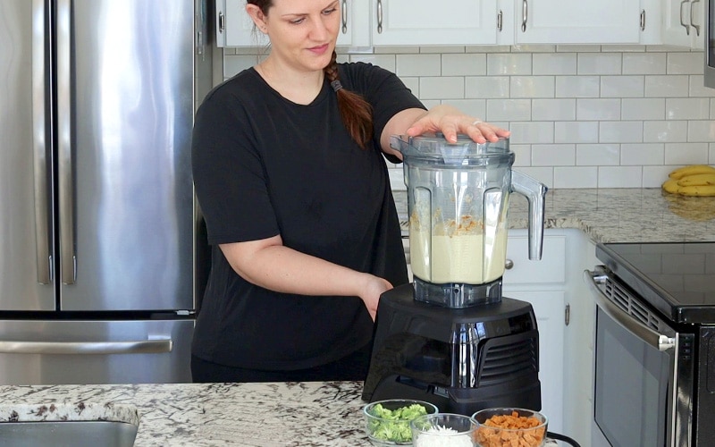 Large blender on counter with creamy beige-colored mixture inside. A women in a black t-shirt stands behind the blender with one hand on the lid and the other on the front of the blender base; she is looking at the blender contents. On the counter in front of the blender are three small glass jars filled with seitan, broccoli and shredded cheese.