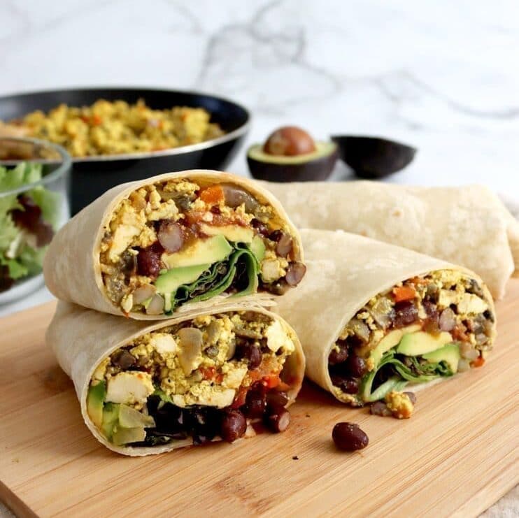 Three burrito halves stacked on wooden board with full burrito in back along with blurred bowl of salad greens, black pan filled with tofu scramble and two avocado halves.