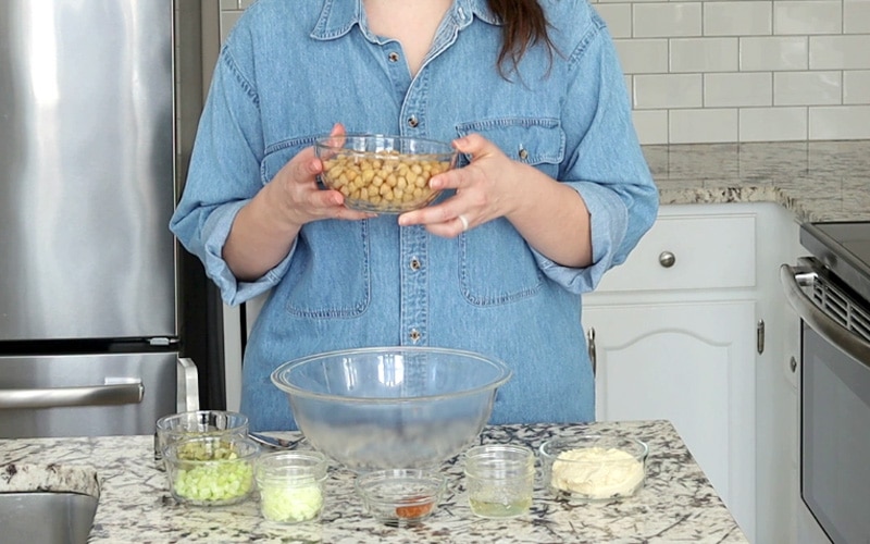 A woman in a denim shirt stands behind a marble counter top, holding a glass bowl full of chickpeas in front of her. Her head is cropped out. On the counter top is a large, empty glass bowl surrounded my smaller glass jars and bowls filled with different ingredients including chopped celery, white onion, pickles, hummus and spices.