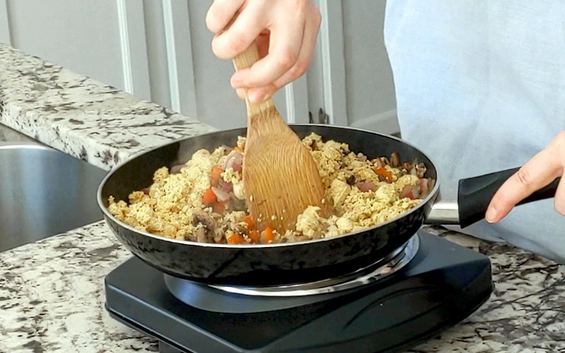 Counter top burner element with large frying pan on top filled with cooked vegetables and crumbled tofu. A hand holding a wood spoon stirs the contents of the pan around.