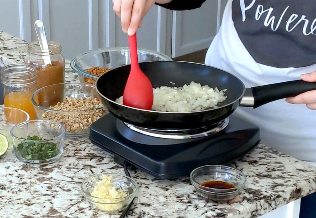A hand holds a large red spoon and is stirring the contents of a black pan filled with chopped onion. Many glass bowls surround the pan on a marble counter top, each filled with a different ingredient (lentils, walnuts, taco spice, vegetable broth, cilantro, garlic, soy sauce).