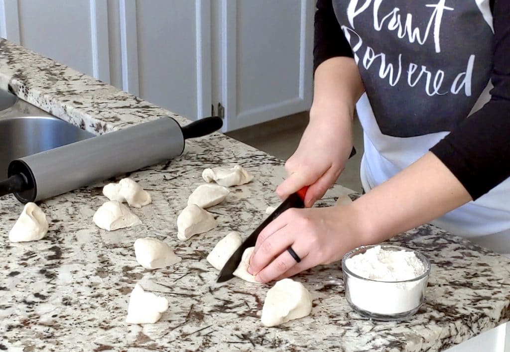A person sliced a piece of dough in half with a large black and red knife on a marble countertop. The counter is covered with scattered pieces of raw dough along with a grey rolling pin and small glass jar of flour.