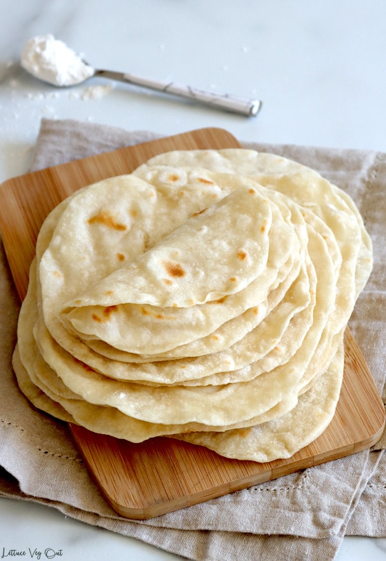Stack of flour tortillas with two folded in half on top of the stack; stack sits on a bamboo board with a light brown towel underneath. A spoon with a scoop of flour sits blurred behind the stack of tortillas.