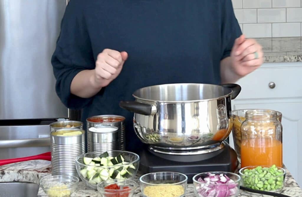 Person stands behind marble counter topped with single burner cooking element with large metal pot on top. This is surrounded by cans of ingredients and other ingredients in small glass bowls or jars.