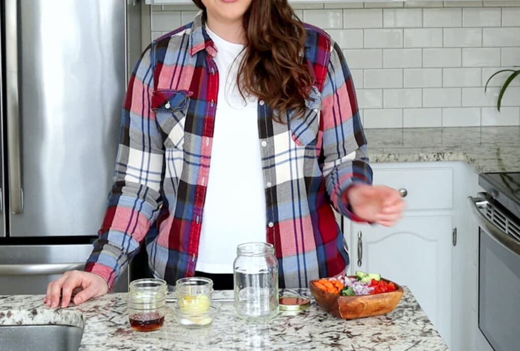 Woman stands behind counter top covered that has an empty mason jar surrounded by smaller jars filled with ingredients to make mustard salad dressing. A garden salad in square wood bowl sits to the right. She wears a red and blue plaid shirt, open with a white shirt underneath.