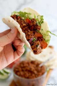 Hand holding up folded flour tortilla filled with cooked walnut lentil taco "meat" with chopped tomato, red onion and shredded lettuce. Blurred in the back is a glass bowl of walnut taco filling and stack of tortillas.