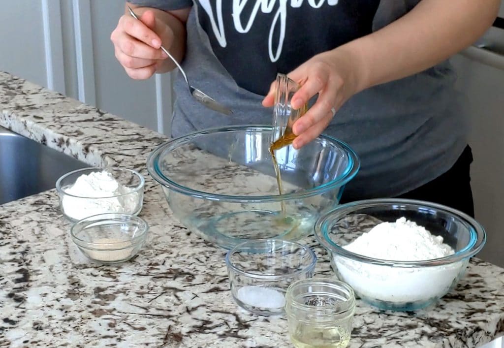 A person stands behind a marble counter with one hand pouring syrup from a small glass dish into a large glass mixing bowl with water at the bottom. The other hand holds a metal spoon over the bowl. Other small glass dishes are placed around the large mixing bowl, each filled with a different ingredient.
