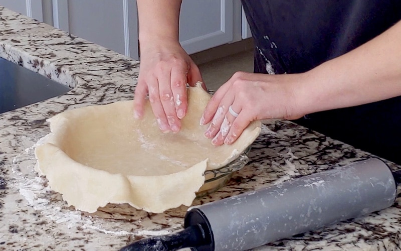Hands press pastry into the edges of a glass pie dish. The marble counter and the hands are covered with white flour and a grey rolling pin sits to the right side of the pie dish.