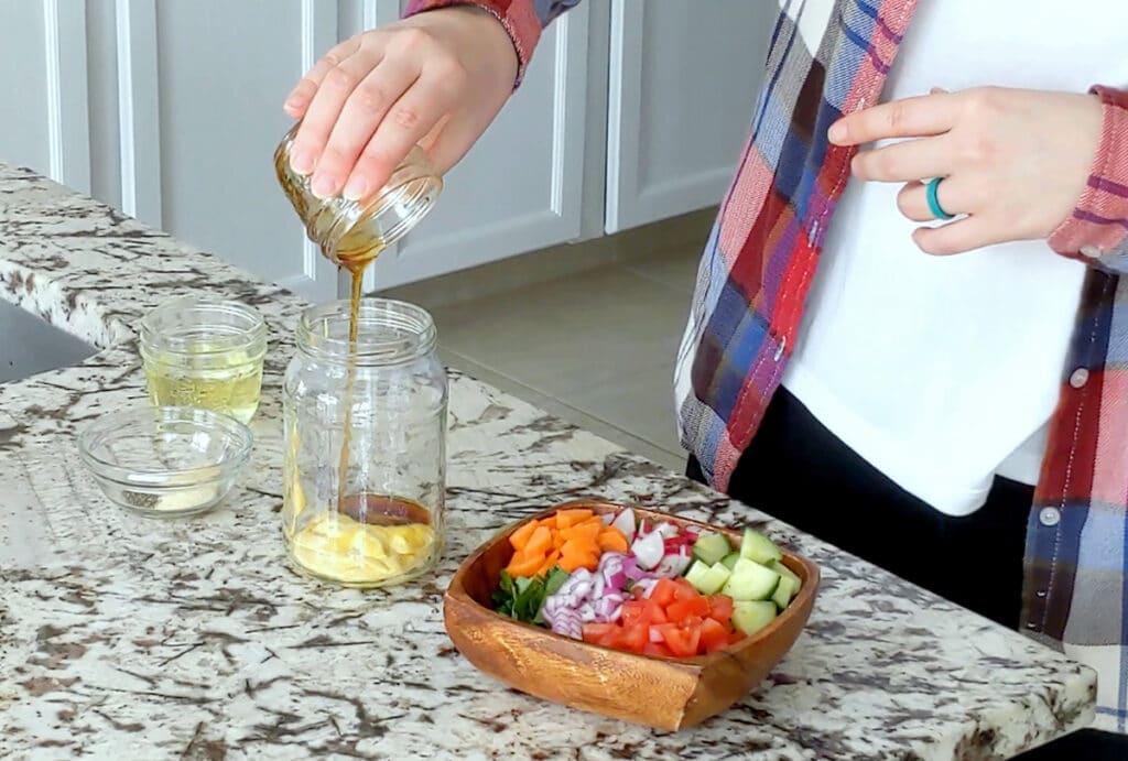 Hand pours maple syrup from a small mason jar into a larger one that contains mustard. Two small glass dishes sit around the larger mason jar on a marble counter top with a square wood bowl filled with garden salad to the right.