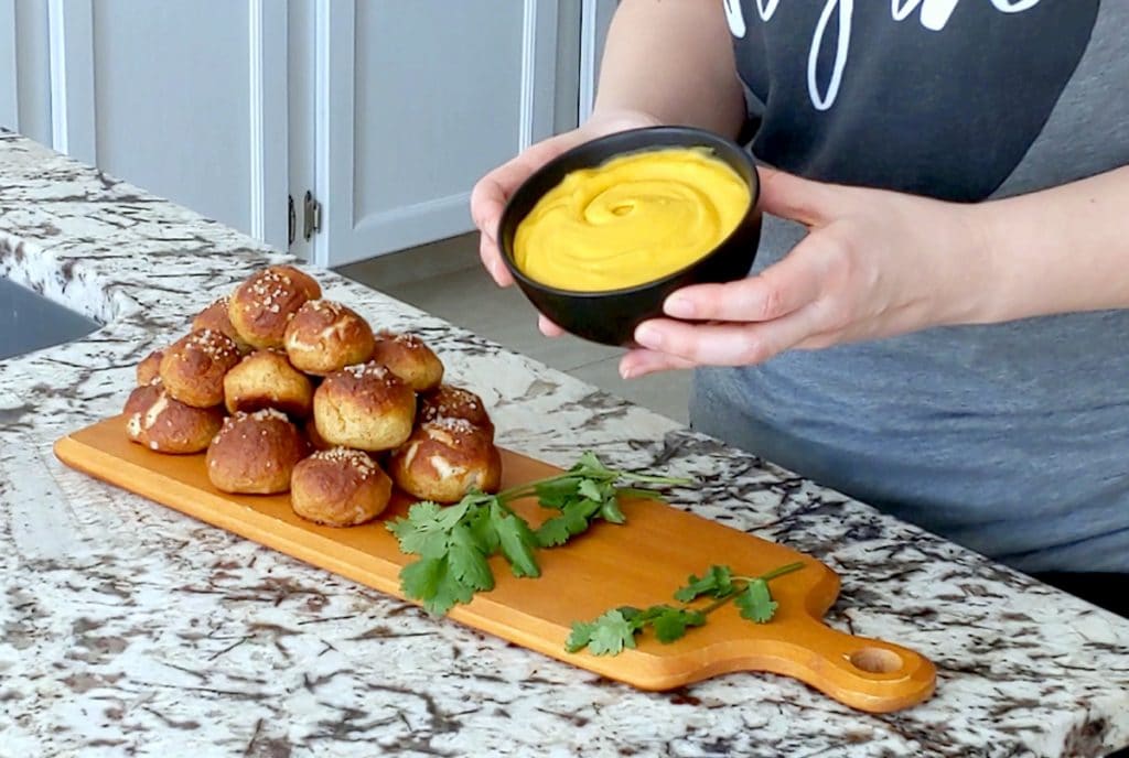 Two hands hold up a small black bowl filled with creamy orange dip. The hands hover over a marble counter top with a long wood board on it, topped with sprigs of cilantro and a stack of baked pretzel bites.