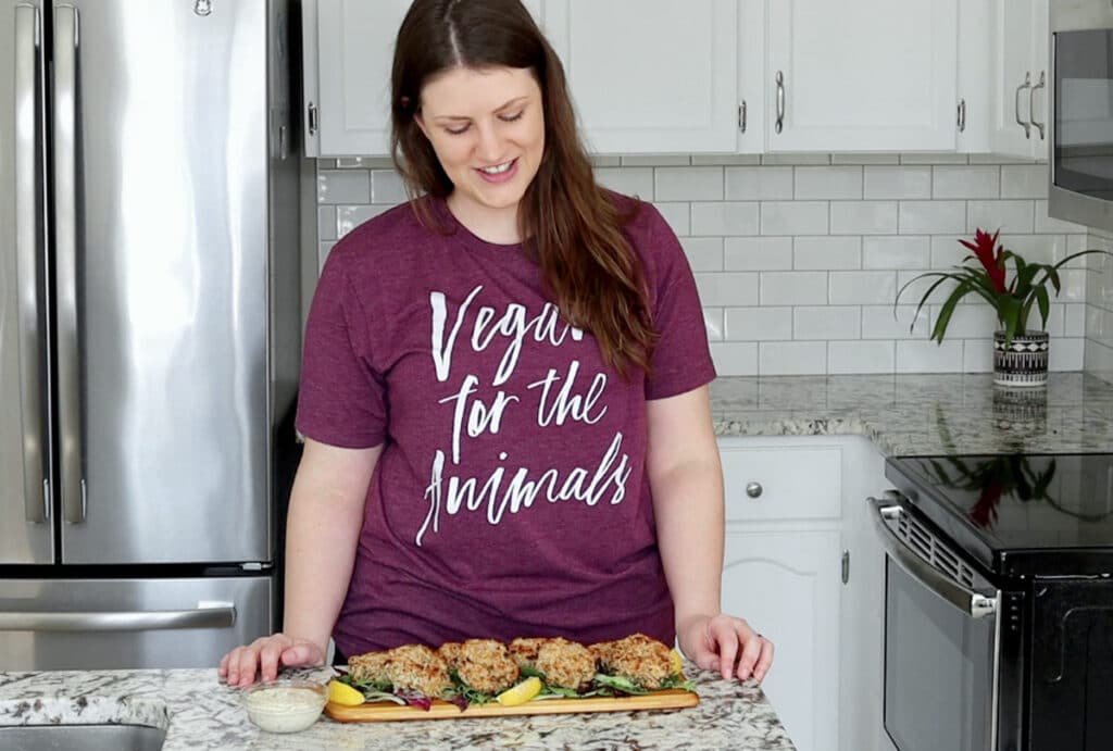 Nicole stands behind a countertop looking down at a narrow wood board topped with salad greens and cooked jackfruit crab cakes with lemon wedges for garnish. She has long brown hair tucked behind her ears and is smiling, with both hands resting on the counter top. She wears a purple t-shirt with white font reading "vegan for the animals"
