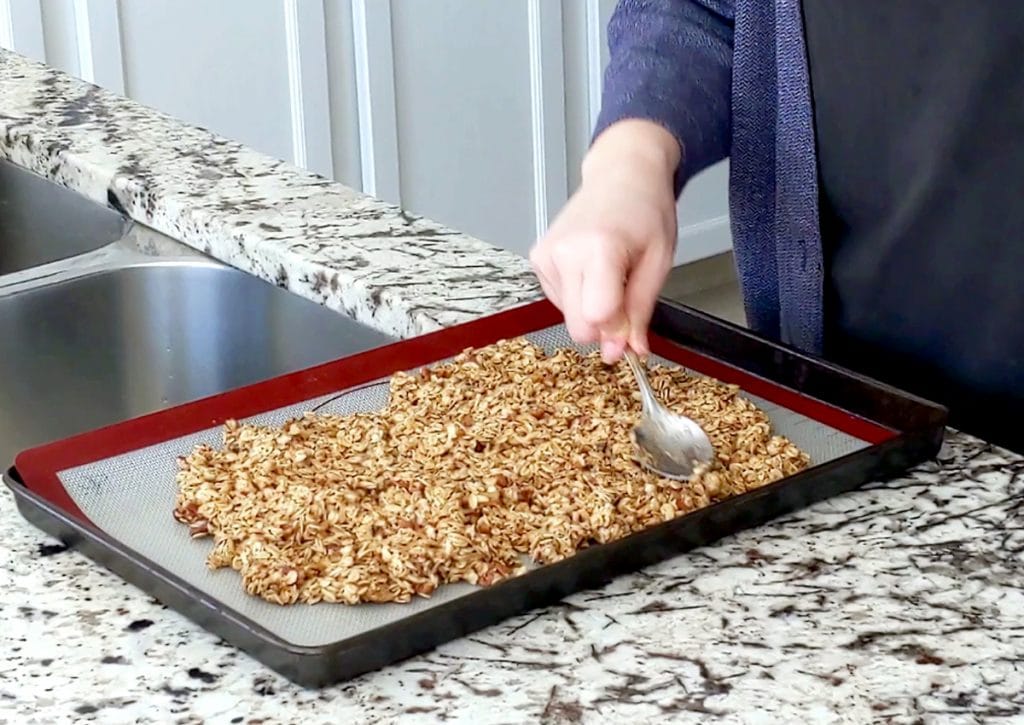 A fair-skinned hand holds a metal spoon and is flattening out a tray full of uncooked granola.