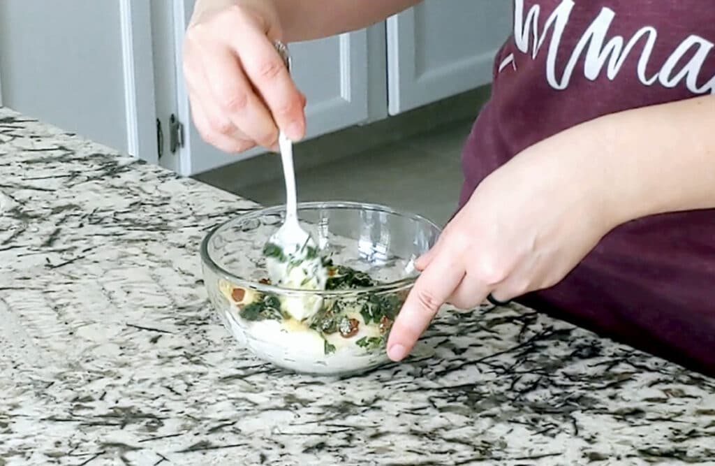 Person mixes a bowl of sour cream dip with chopped parsley and hot sauce visible. One hand holds the edge of the bowl and the other a metal spoon.