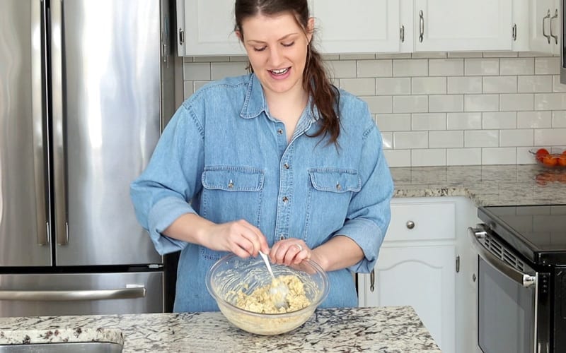 A light skinned woman stands behind a marble counter top. She is stirring chickpea salad in a large glass bowl and her gaze is down on the bowl. She wears a large blue denim shirt and has long wavy brown hair tied in a low ponytail.
