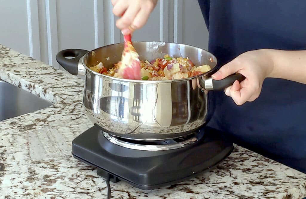Stirring ingredients for red curry pasta in large metal pot on single cooking element set on marble counter. One of the person's hands holds the handle of the pot while the other holds a large red spoon and is slightly blurred in motin.