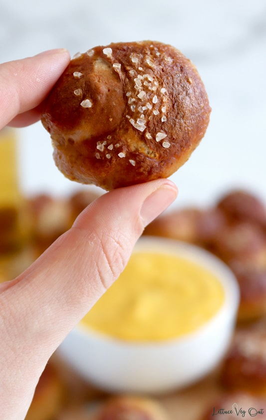 Hand holding up a round, baked soft pretzel bite that is dark brown in color and topped with salt. Background is blurred but shows a white bowl filled with creamy orange dip, surrounded by other soft pretzel bites.