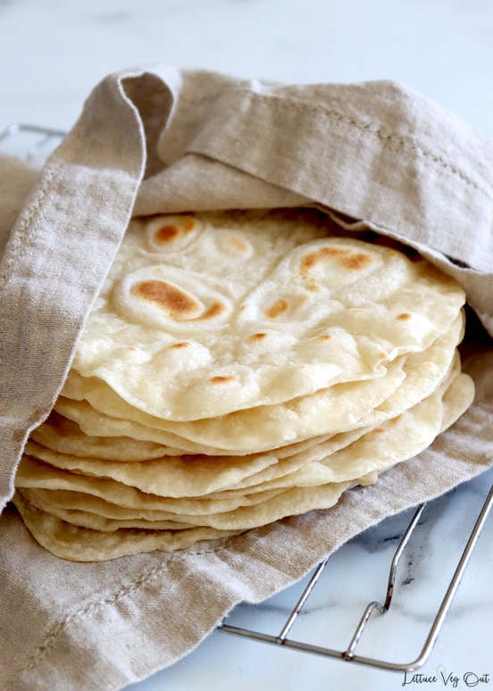 A stack of vegan tortillas sit wrapped in a light brown towel over top a metal wire cooling rack on a marble counter.