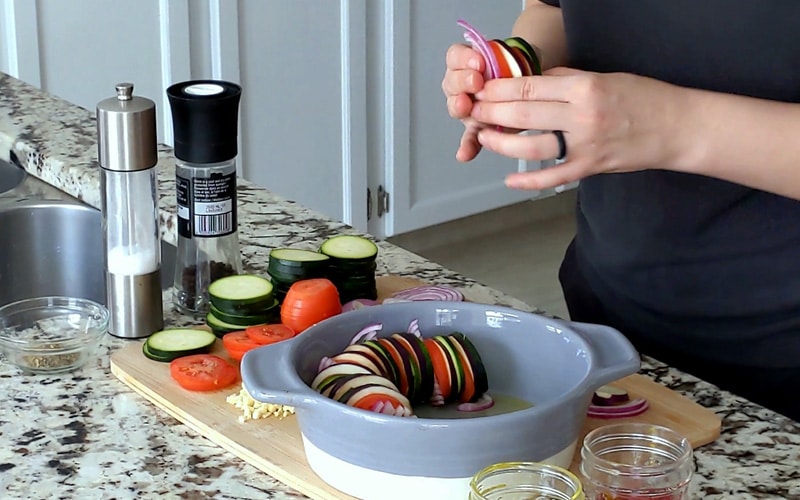 Hands holding a row of sliced vegetables over a round, grey ceramic baking dish half full of sliced vegetables sitting on their edges (tomato, zucchini, eggplant and red onion). Sliced vegetables and other ingredients surround baking dish on marble counter.