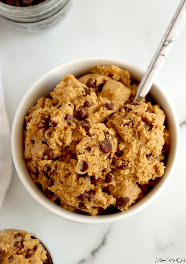 Top view of white bowl filled with raw chickpea cookie dough with mini chocolate chips. Marble spoon in cookie dough, with a scoop of cookie dough in bottom left corner of image. White-grey marble background.