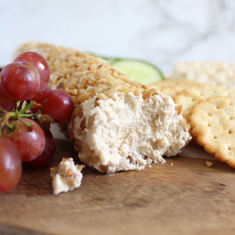 Vegan goat cheese log coated with crushed walnuts, on wooden board surrounded with crackers, red grapes and cucumber slices