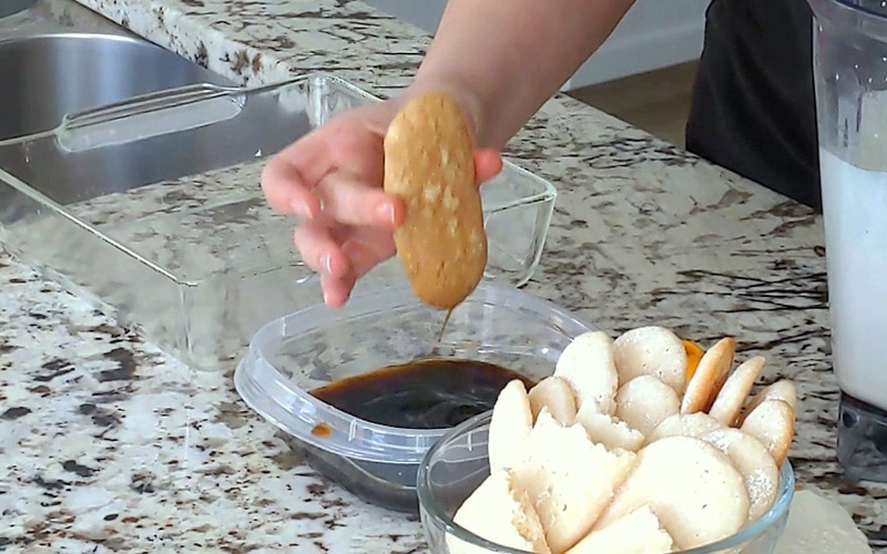 Hand holding a dark brown ladyfinger cookie between the thumb and middle finger. A drip of coffee falls from the cookie into a plastic container of coffee below. Glass bowl of light-colored ladyfingers to the right with empty glass dish to the left.